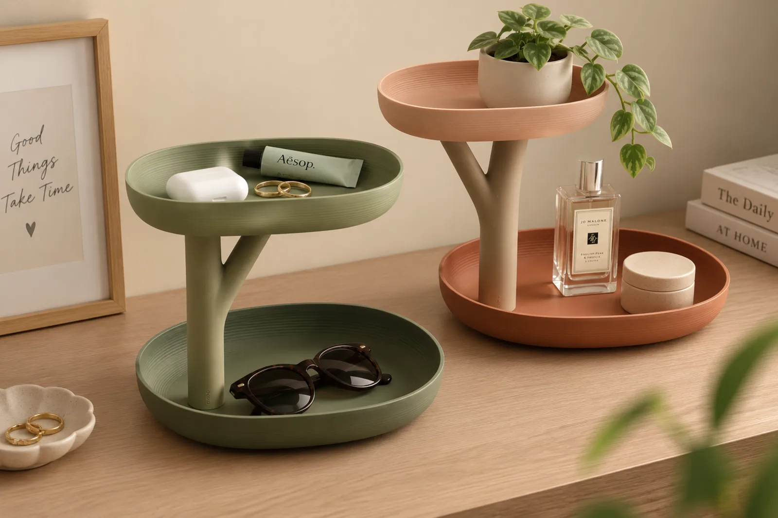 Dos organizadores de dos niveles sobre una cómoda, uno verde olivo y otro terracota, con lentes, frascos y una planta encima.